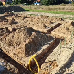 Wykop pod fundamenty z ułożonym zbrojeniem i żółtą rurą instalacyjną w rogu, widok z góry na tle zabudowań wiejskich i zieleni.