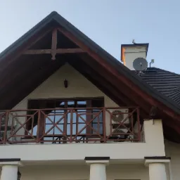 Elewacja domu w Rykach z balkonem, na którym zamontowano dwie jednostki zewnętrzne klimatyzacji, widoczne spod dachu z dekoracyjnym belkowaniem i oknem w kształcie oka.