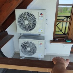 Jarosław Buczek ACTUAL COMFORT - Dwa klimatyzatory marki Gree zamontowane na białej ścianie zewnętrznej domu z drewnianą konstrukcją dachu i oknem w tle.