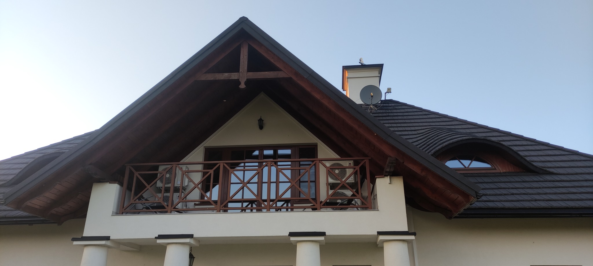 Elewacja domu w Rykach z balkonem, na którym zamontowano dwie jednostki zewnętrzne klimatyzacji, widoczne spod dachu z dekoracyjnym belkowaniem i oknem w kształcie oka.