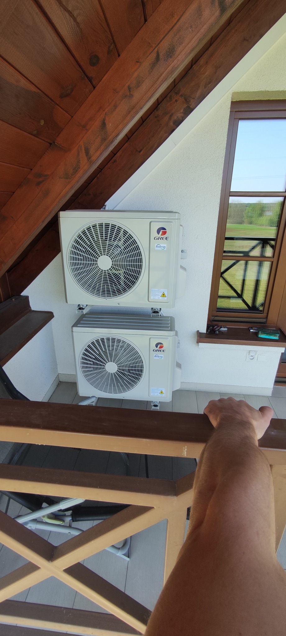 Dwa klimatyzatory marki Gree zamontowane na białej ścianie zewnętrznej domu z drewnianą konstrukcją dachu i oknem w tle.