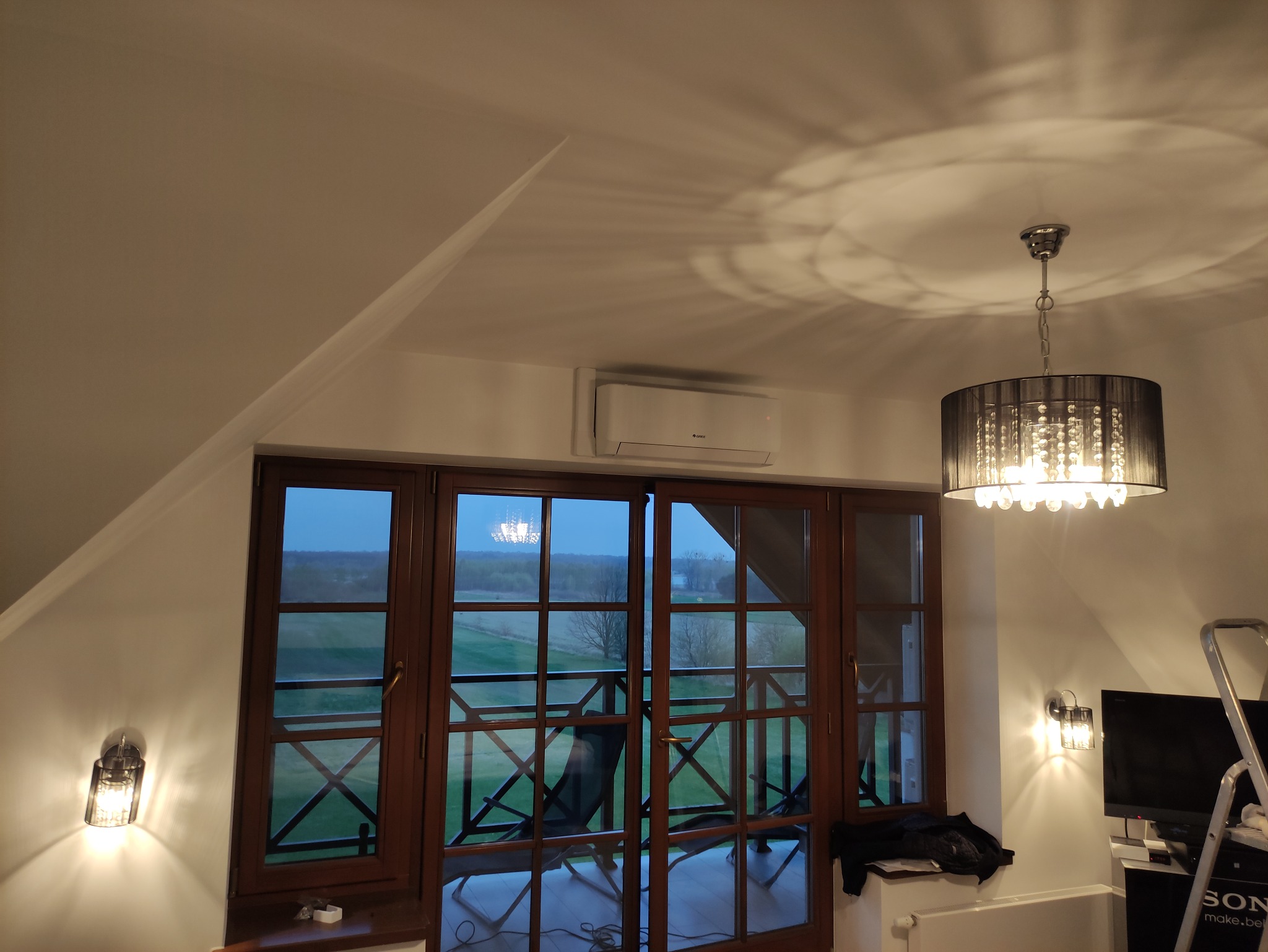 Wnętrze poddasza z zamontowanym klimatyzatorem typu split, widok na zewnątrz przez drewniane okno balkonowe, ozdobny żyrandol i kinkiet.