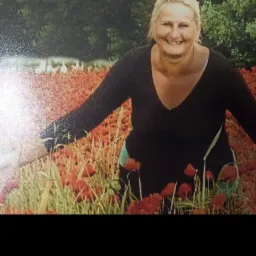 Uśmiechnięta niania w czarnej bluzce, stojąca w polu czerwonych maków z rozłożonymi ramionami, na tle zielonych drzew.