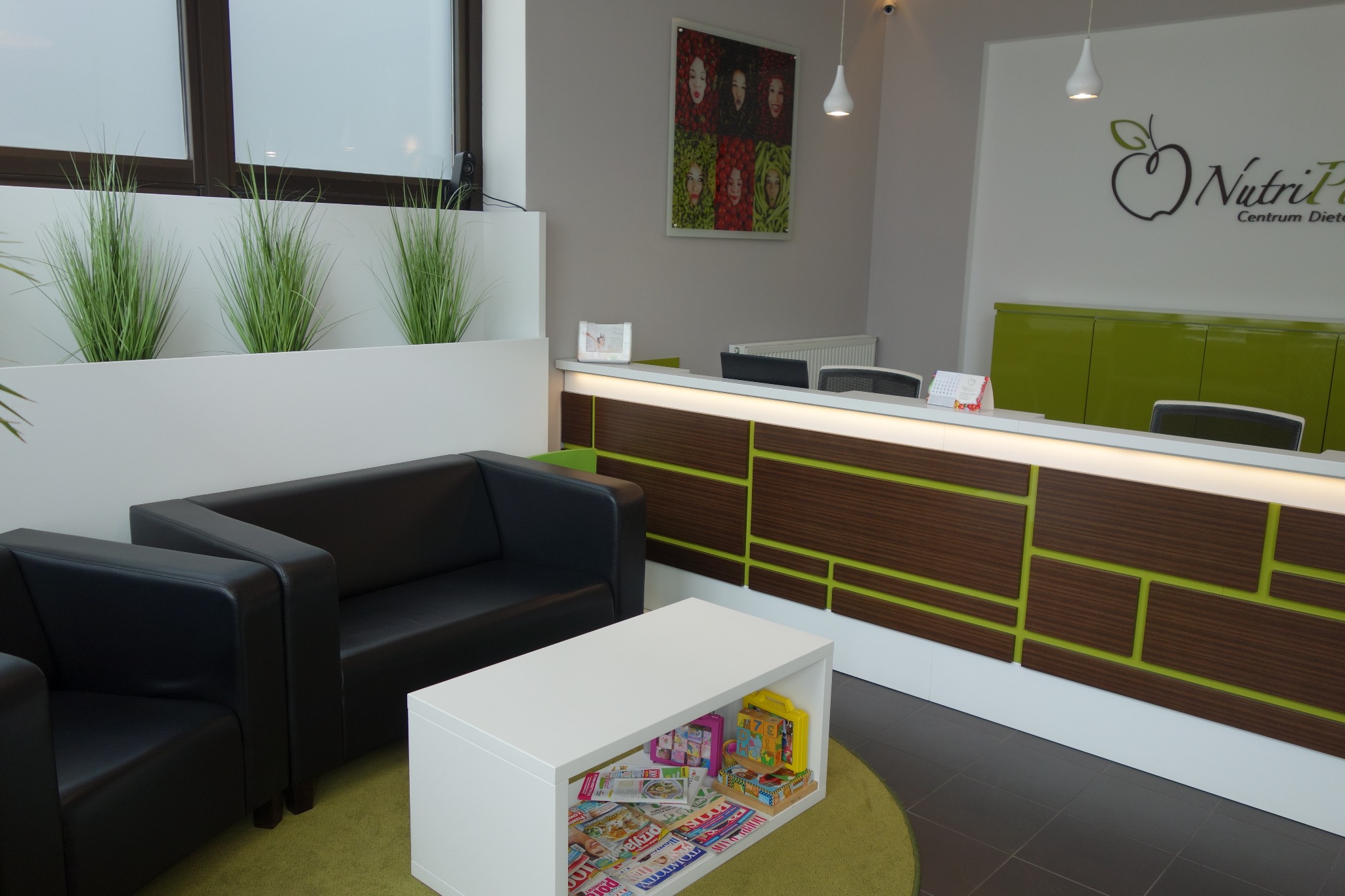 Wnętrze poczekalni centrum dietetycznego z recepcją, sofami i stolikiem kawowym z magazynami. Na ścianie logo centrum i grafika z portretami. Rośliny w donicach.