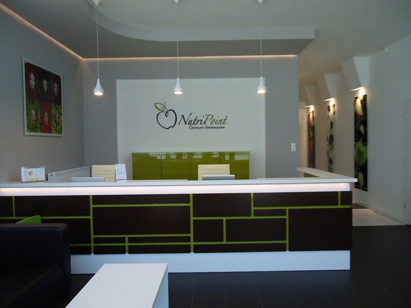 Recepcja w gabinecie poradnictwa żywieniowego NutriPoint, z ciemnobrązowymi i zielonymi elementami dekoracyjnymi, widoczne logo na ścianie i grafiki z portretami osób i warzyw.
