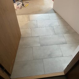 Adrian Gęstwicki - Świeżo ułożone, podłużne, szare płytki podłogowe w przejściu, widoczne przez otwarte drewniane drzwi. W tle wiadro, puszki i kartony z materiałami budowlanymi.