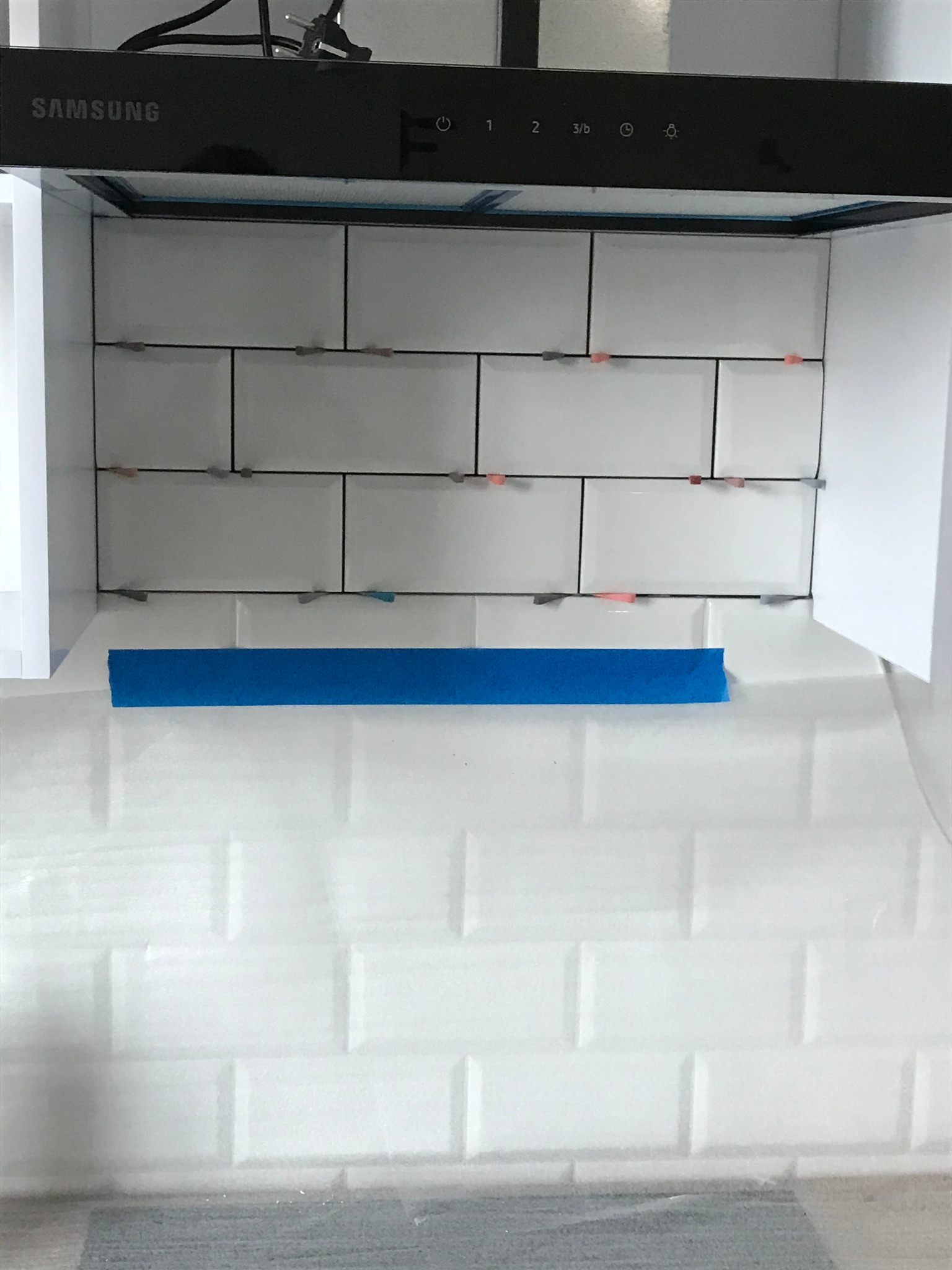 Białe, prostokątne płytki ułożone na ścianie w kuchni pod okapem Samsung, z widocznymi krzyżykami dystansowymi i niebieską taśmą zabezpieczającą.