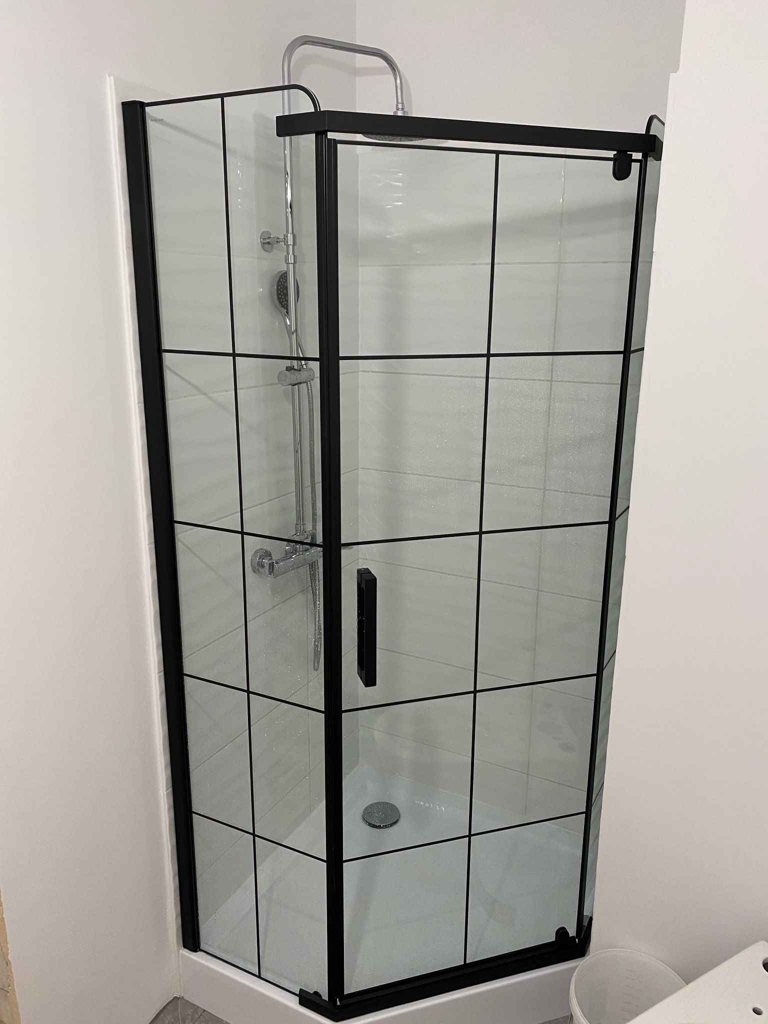 Kabina prysznicowa z czarnymi profilami i szklanymi ściankami w kratkę, zamontowana w narożniku wykończonej łazienki z białymi płytkami.