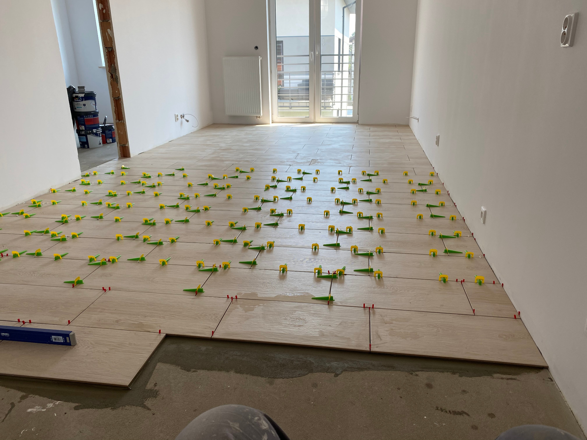 Jasne płytki podłogowe imitujące drewno, ułożone w dużym pokoju z widocznymi klipsami poziomującymi i krzyżykami dystansowymi, częściowo na surowym betonie, z oknem balkonowym w tle.