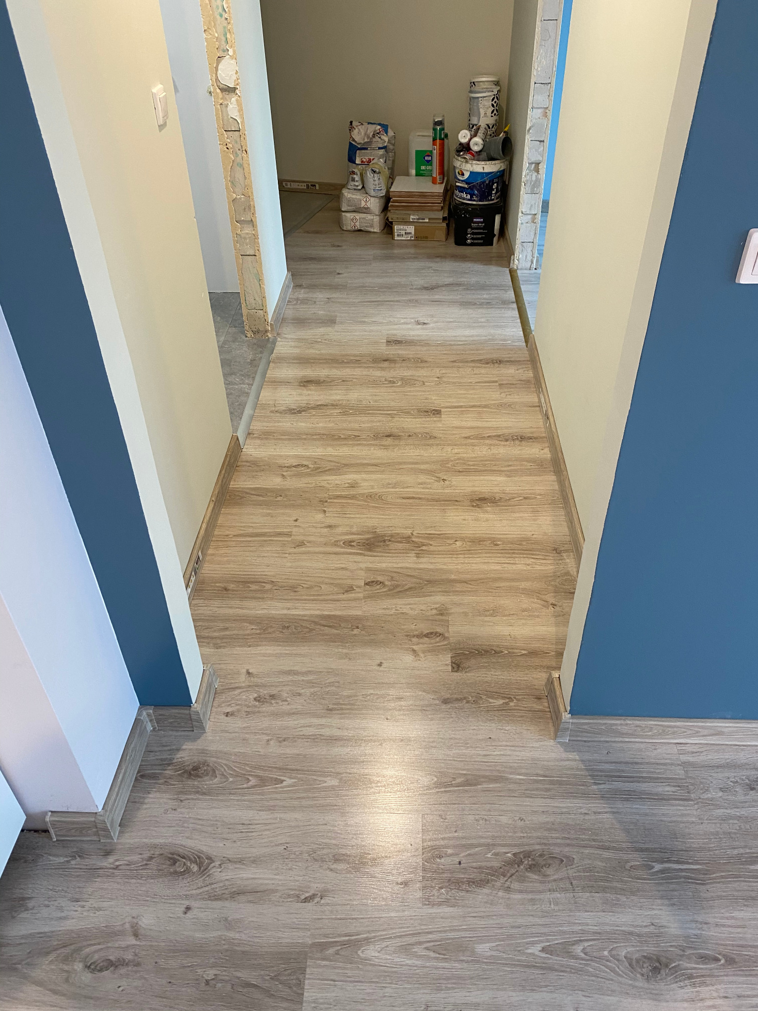 Korytarz z nową podłogą z paneli imitujących drewno, ściany w odcieniach bieli i granatu, widoczne puszki z farbą i materiały budowlane w tle, sugerujące trwający remont.