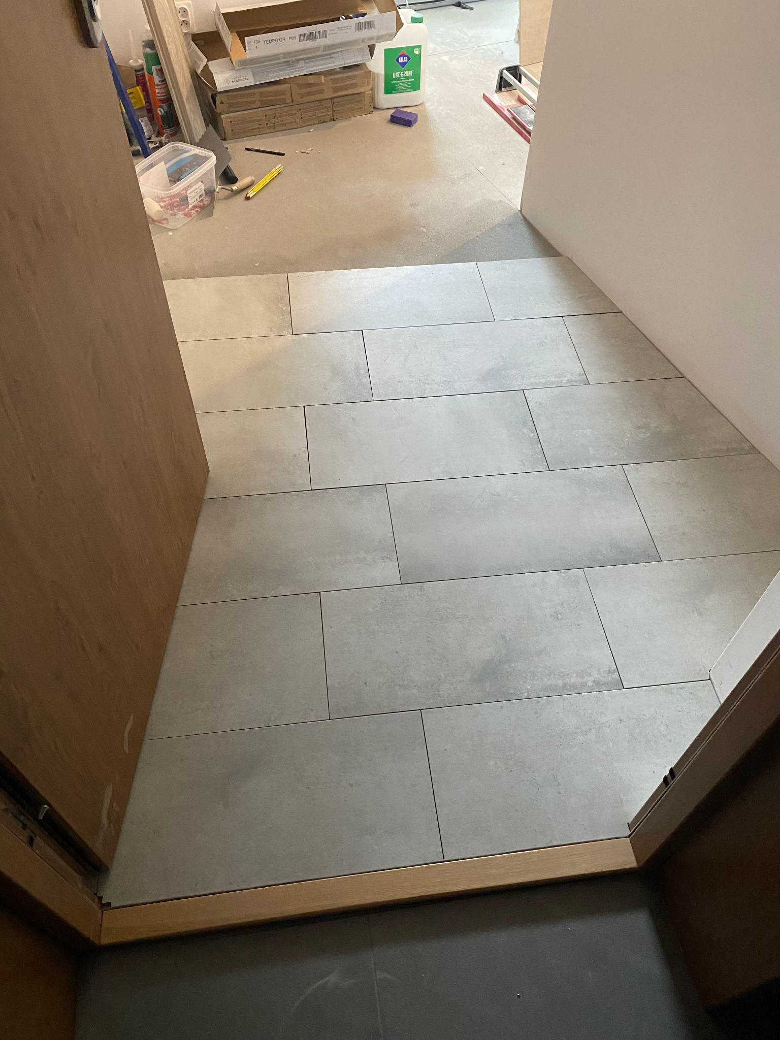 Świeżo ułożone, podłużne, szare płytki podłogowe w przejściu, widoczne przez otwarte drewniane drzwi. W tle wiadro, puszki i kartony z materiałami budowlanymi.