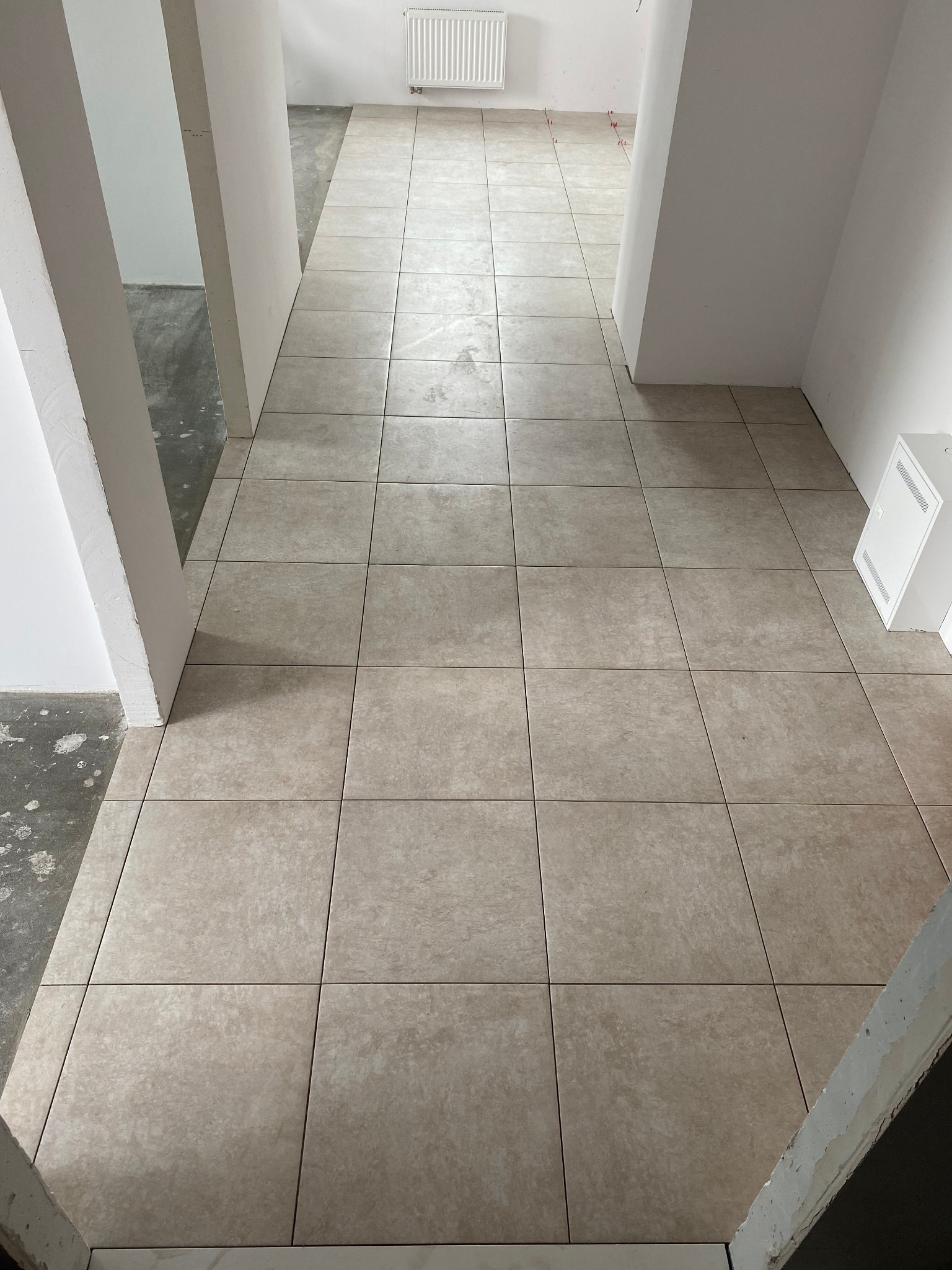 Korytarz z ułożoną beżową terakotą, widoczne krzyżyki dystansowe, surowy beton na ścianach w tle, białe ściany i grzejnik.