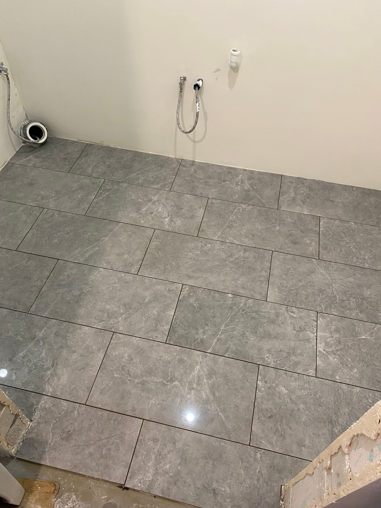 Ułożone szare, prostokątne płytki imitujące marmur na podłodze łazienki, widoczne przyłącza wodne na ścianie i niedokończone wykończenie przy progu.
