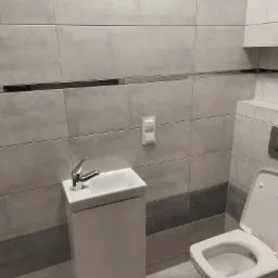 Nowoczesna łazienka z umywalką nablatową na szafce i podwieszaną toaletą, wykończona szarymi płytkami z dekoracyjnym pasem.