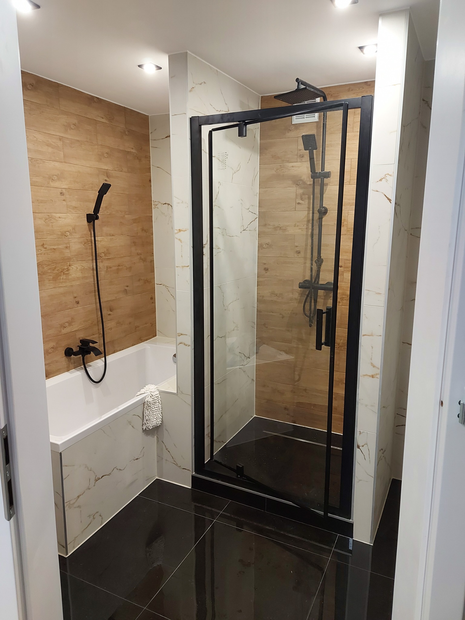 Nowoczesna łazienka z kabiną prysznicową typu walk-in z czarnymi ramami, wanną obudowaną marmurowymi płytami, armaturą w kolorze czarnym i drewnopodobnymi panelami na ścianie.