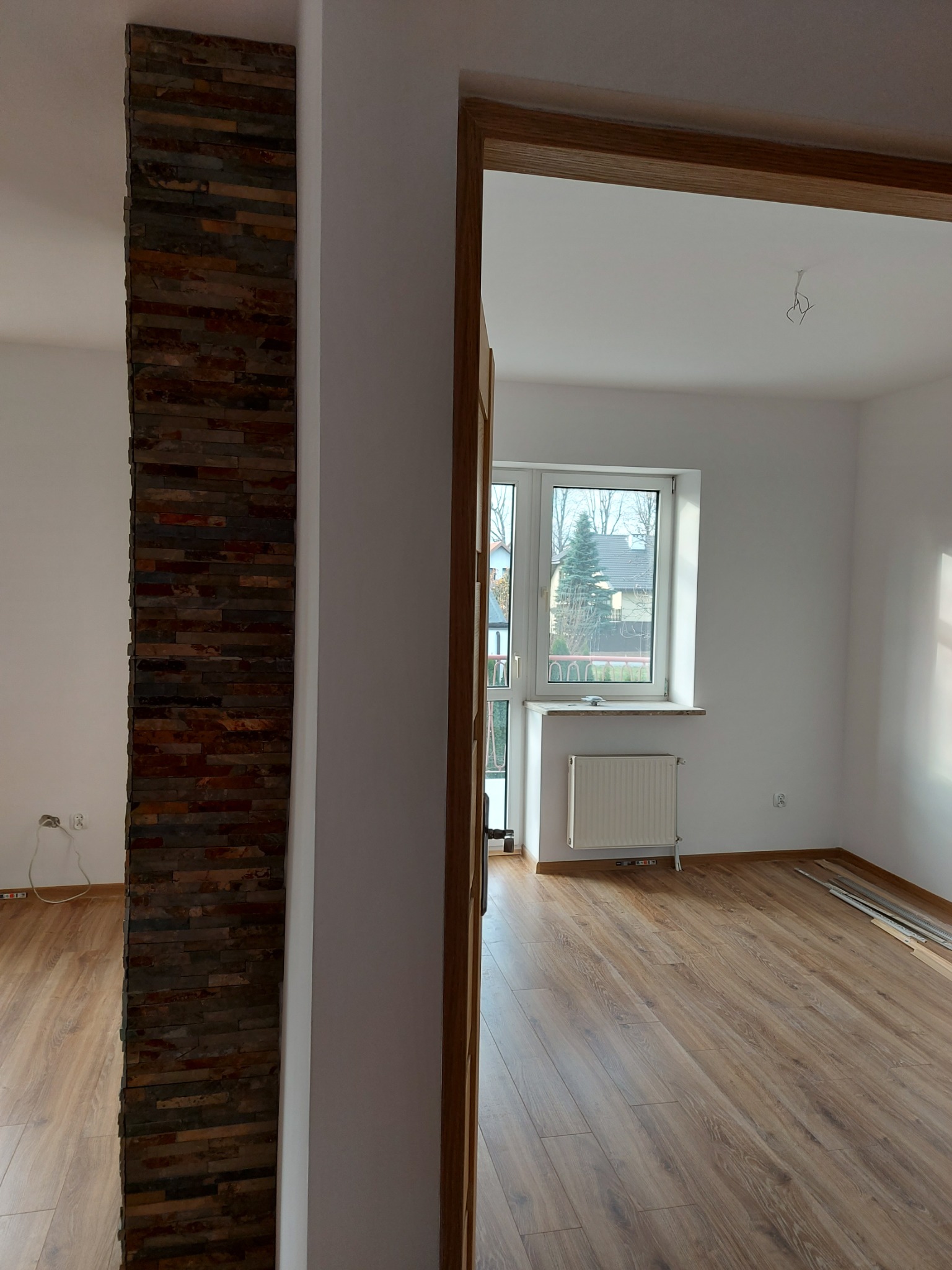 Wnętrze pokoju po remoncie: drewnopodobne panele podłogowe, fragment ściany wykończony dekoracyjnym kamieniem, widoczne listwy przypodłogowe i okno z grzejnikiem.
