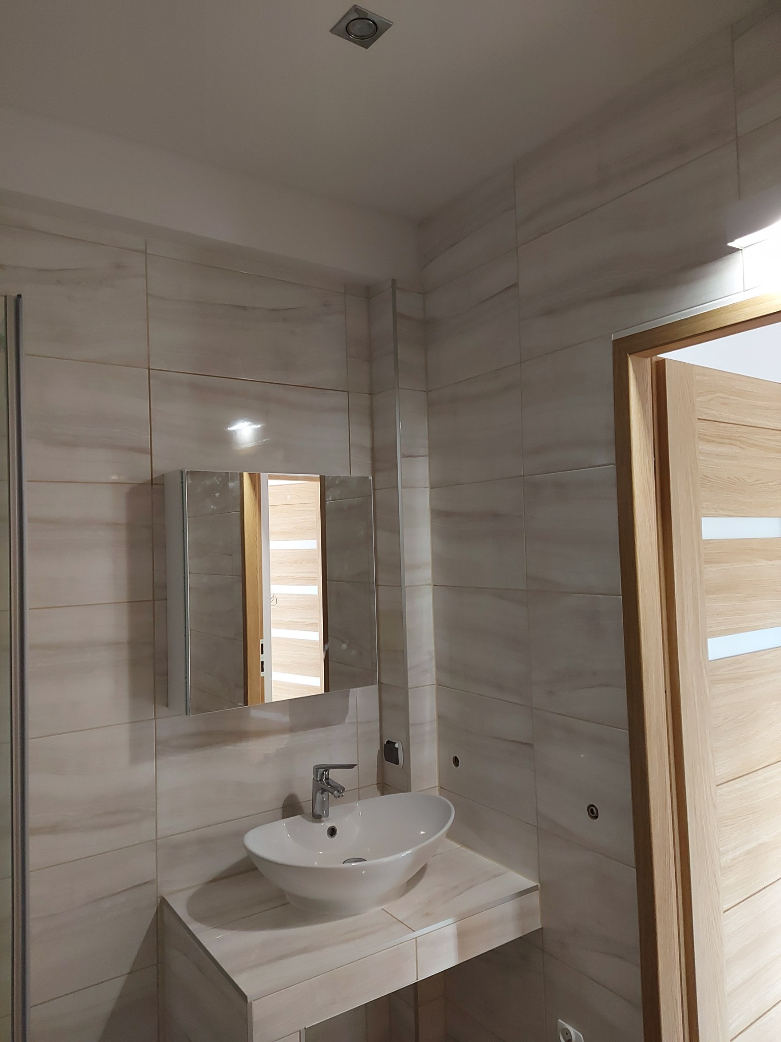 Nowoczesna łazienka z umywalką nablatową, lustrem w szafce i płytkami imitującymi marmur na ścianach, widoczny fragment drzwi z jasnego drewna.