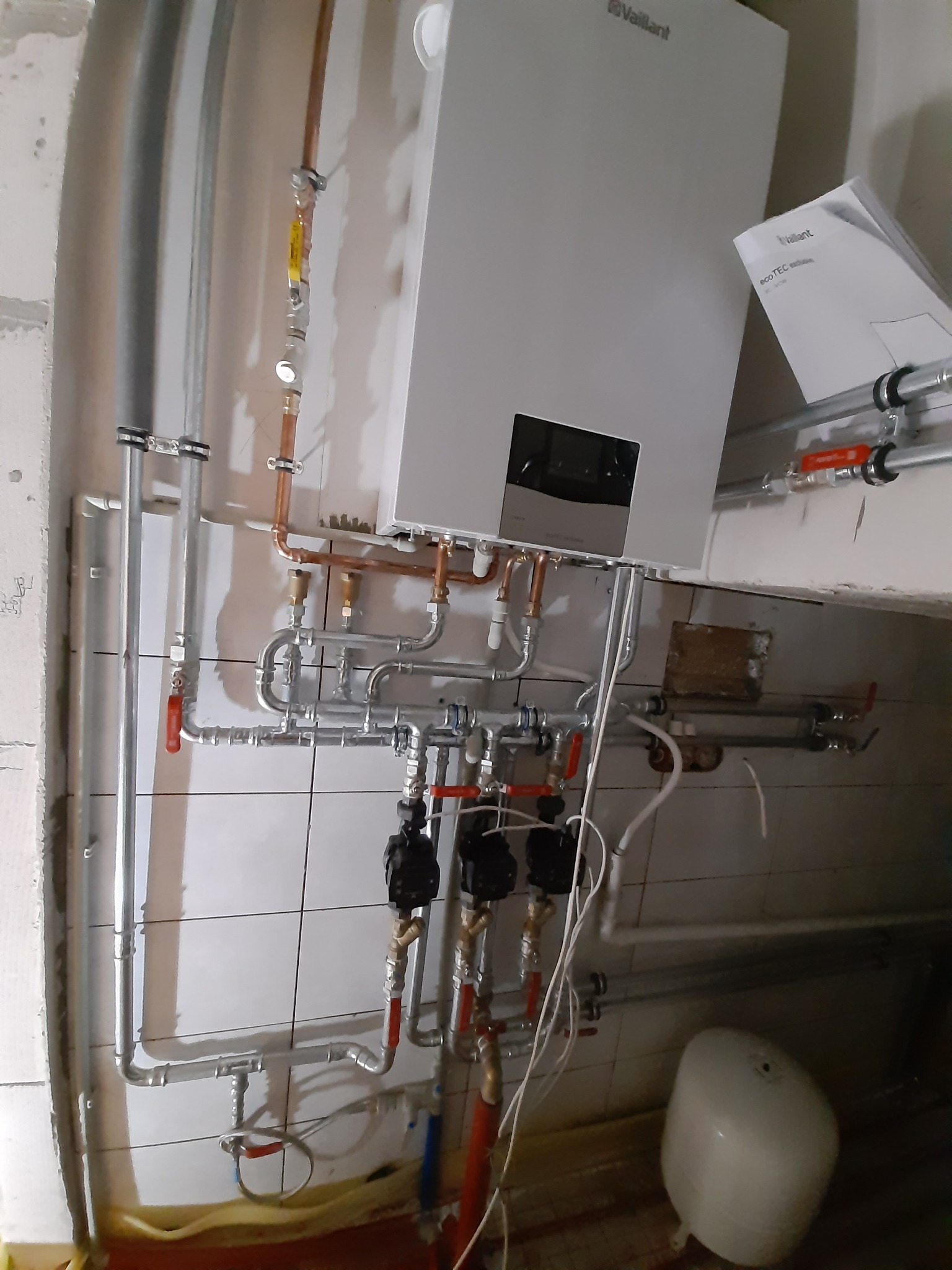 Montaż kotła gazowego Vaillant z widocznymi rurami miedzianymi i zaworami na tle białych płytek w surowym stanie.
