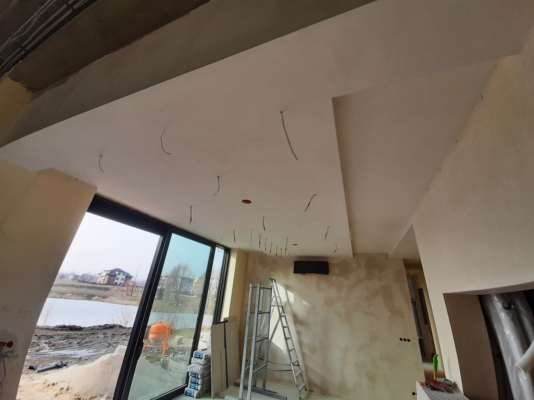 Wnętrze w trakcie wykończenia: gładkie ściany, podwieszany sufit z wystającymi kablami, drabina aluminiowa, widok na jezioro przez duże okno, worki z gipsem przy ścianie.