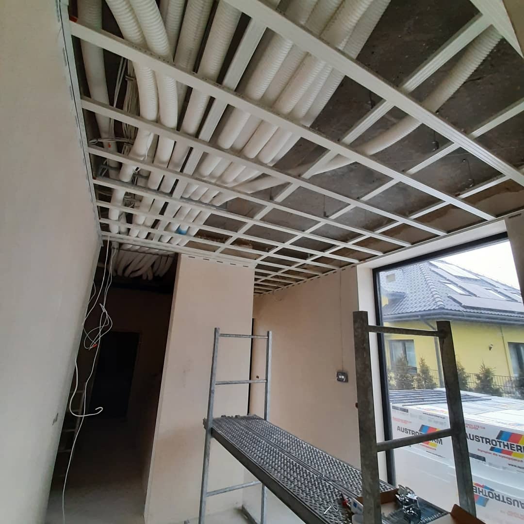 Metalowa konstrukcja sufitu podwieszanego z widocznymi rurami wentylacyjnymi, drabina aluminiowa w pomieszczeniu w trakcie prac wykończeniowych.