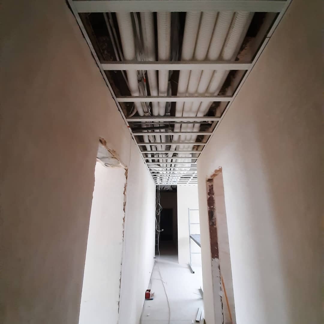 Korytarz w trakcie montażu sufitu podwieszanego z widocznymi instalacjami elektrycznymi i wentylacyjnymi, ściany przygotowane do malowania.