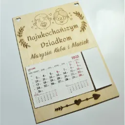 Drewniany kalendarz z wygrawerowanym napisem 'Najukochańszym Dziadkom' oraz imionami Marysia, Kuba i Maciek, ozdobiony rysunkami dziadka, babci i serduszek, z widocznym kalendarium na styczeń...