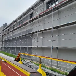Szara ściana budynku w trakcie prac elewacyjnych, osłonięta rusztowaniem, widoczna z perspektywy bieżni sportowej z przeszkodami w pierwszym planie.
