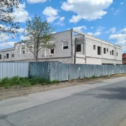 Surowy stan dwukondygnacyjnego budynku z białych bloczków, ogrodzony metalowym płotem tymczasowym, widok z ulicy w słoneczny dzień.