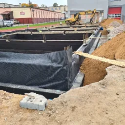 Wykop pod fundamenty z izolacją przeciwwilgociową i deskowaniem, hałda piasku i pracujący koparka w tle.