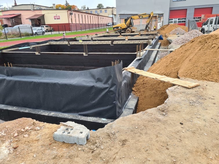 Wykop pod fundamenty z izolacją przeciwwilgociową i deskowaniem, hałda piasku i pracujący koparka w tle.