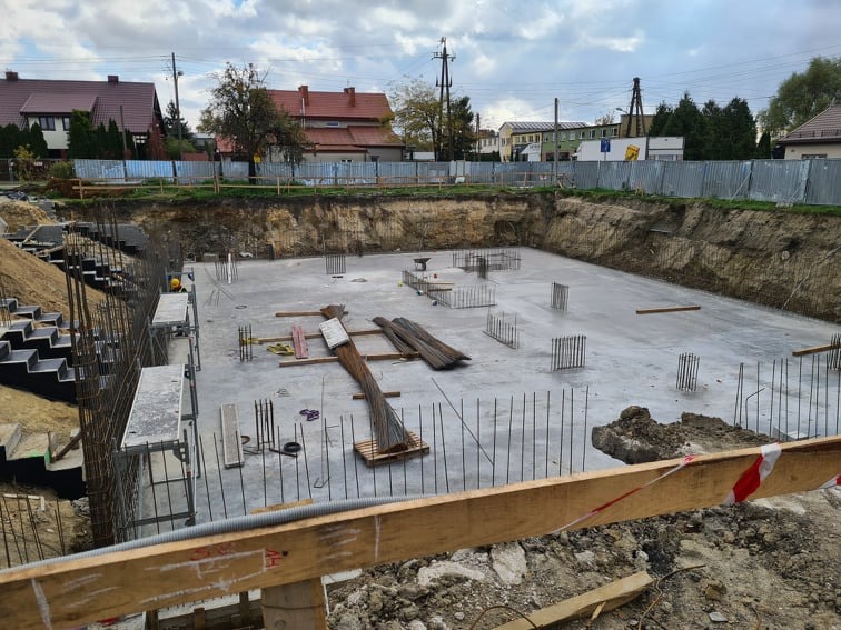 Przygotowany fundament z wystającymi prętami zbrojeniowymi, widok z góry na wykop, w tle zabudowa jednorodzinna.