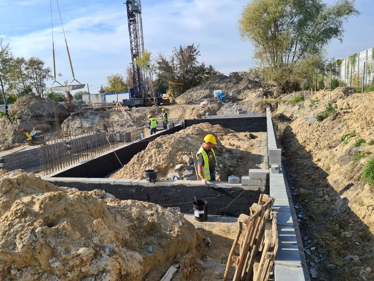 Budowa fundamentów: pracownicy w żółtych kaskach murowują ściany z betonowych bloczków, widoczny dźwig podnoszący element konstrukcyjny, grunt zabezpieczony czarną izolacją.