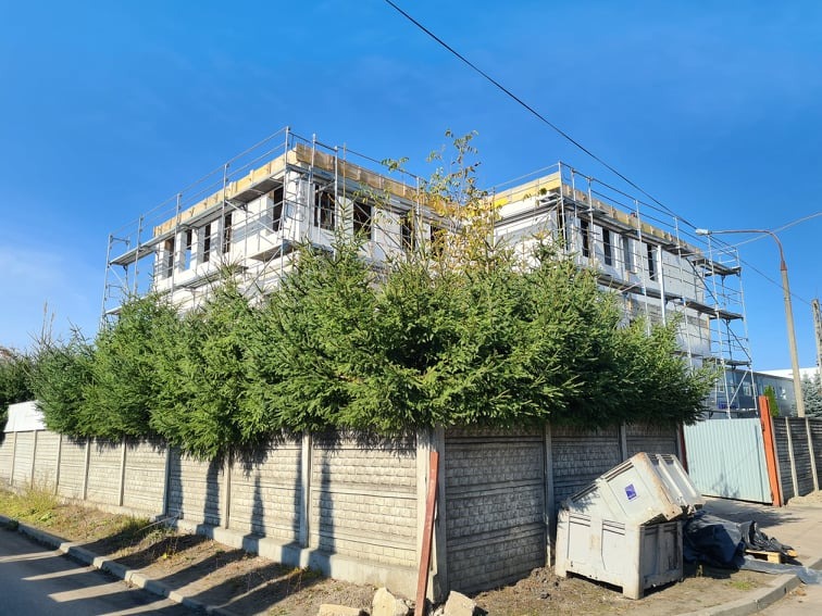 Dwupiętrowy budynek w trakcie ocieplania elewacji, widoczne rusztowania, styropian na ścianach, zieleń i betonowe ogrodzenie na pierwszym planie.