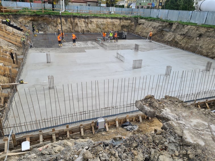 Wylewanie betonu na zbrojenie fundamentu domu jednorodzinnego, widoczni pracownicy w odblaskowych kamizelkach, szalunki i wystające pręty zbrojeniowe.