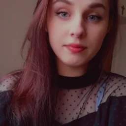 Portret młodej kobiety z różowymi włosami i niebieskimi oczami, ubrana w czarną bluzkę w kropki, delikatny makijaż i subtelny uśmiech.