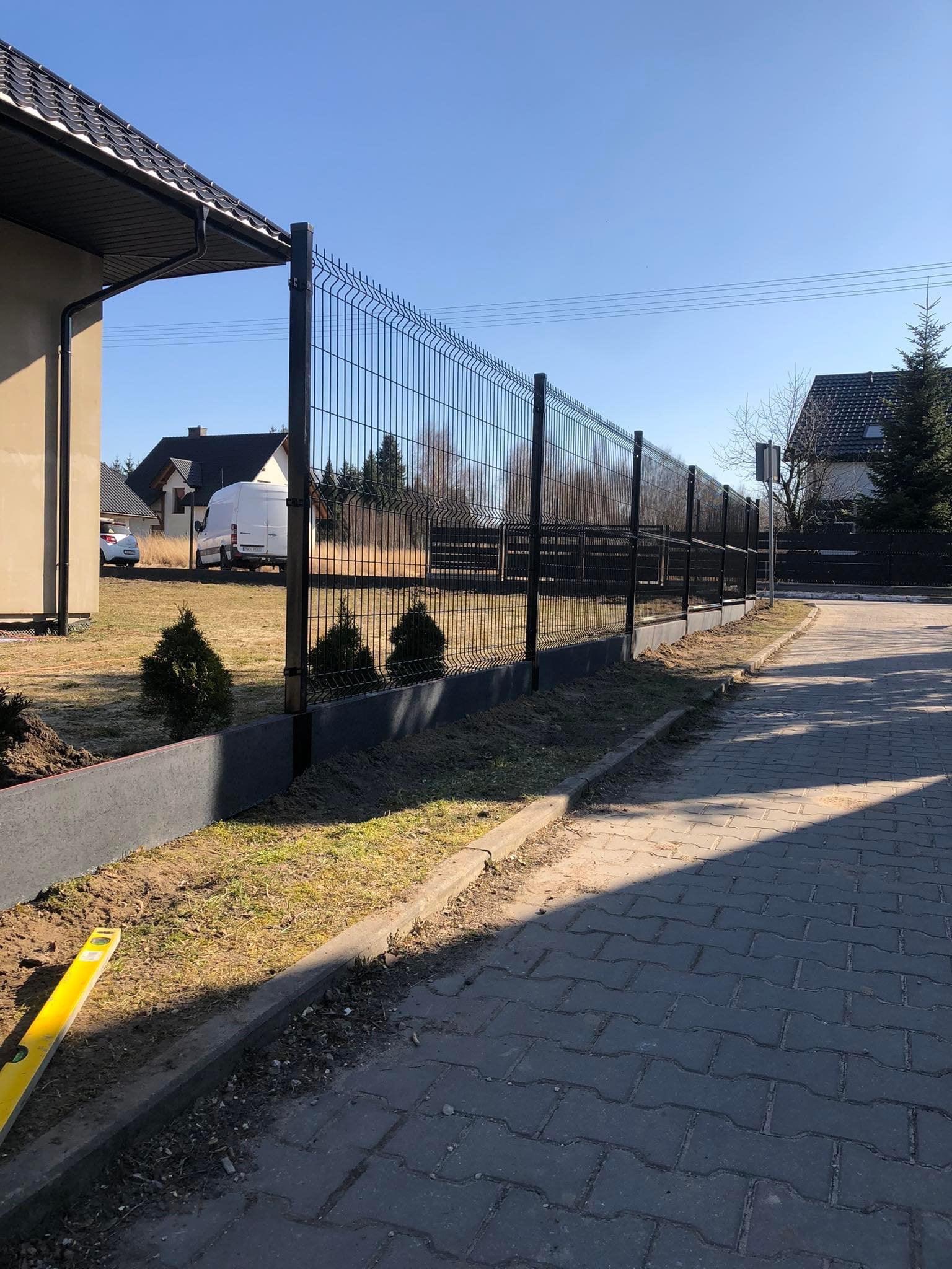 Nowoczesne ogrodzenie panelowe w kolorze grafitowym z podmurówką, widok z zewnątrz posesji, w tle domy i samochód dostawczy, poziomica leżąca na ziemi.