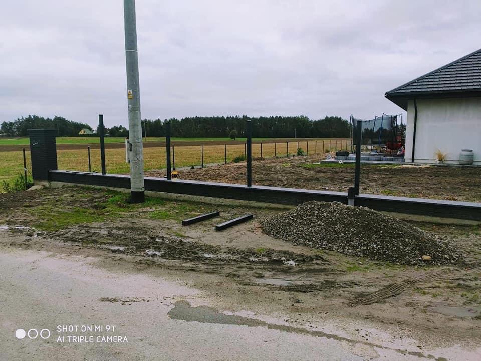 Montaż ogrodzenia panelowego z betonową podmurówką w trakcie realizacji: widoczne słupki, podmurówka, nasyp kamieni i teren budowy z domem w tle.