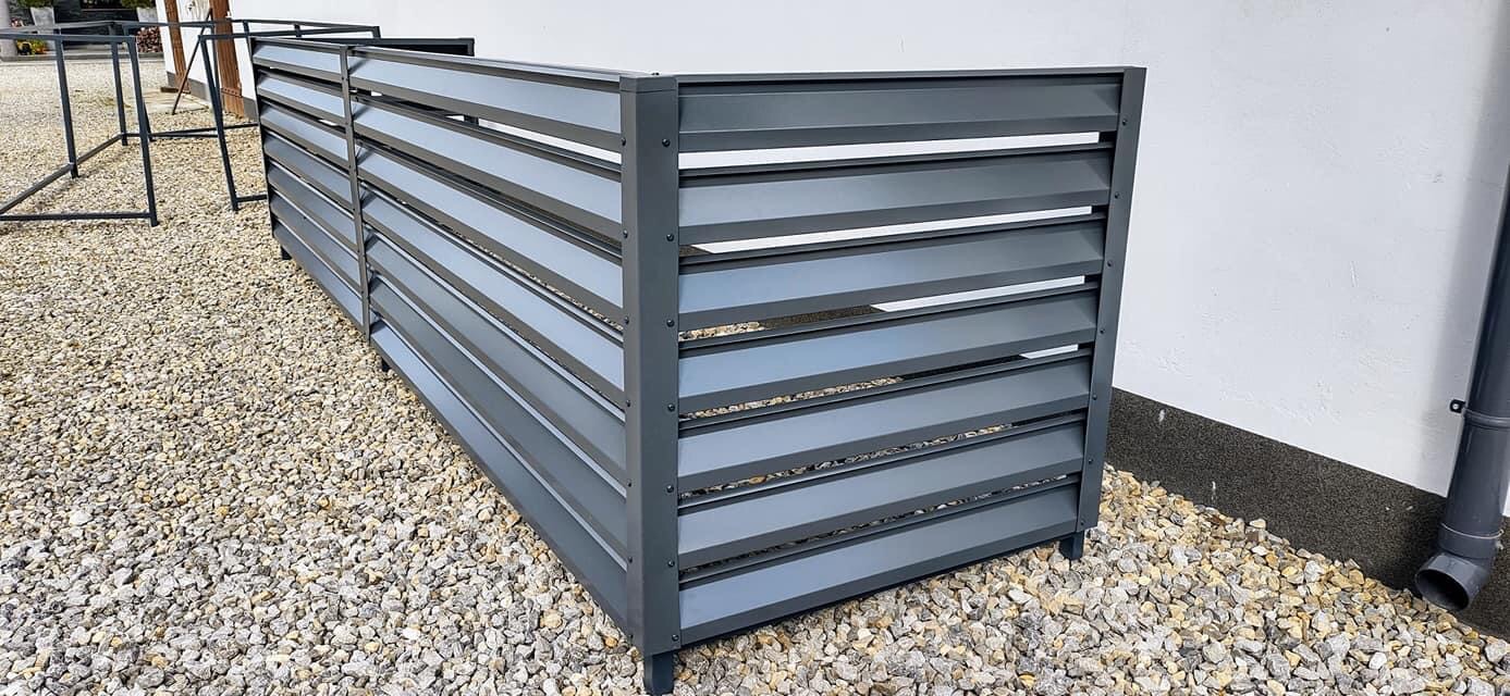 Szare, metalowe przesłony ogrodowe z poziomymi lamelami, ustawione na żwirowym podłożu przy białej ścianie budynku.