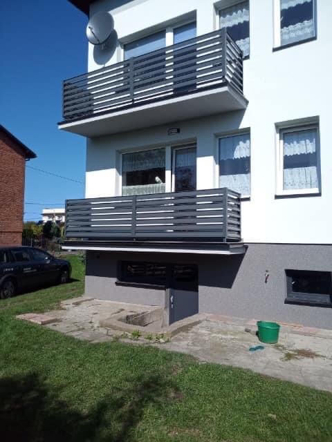 Szare, poziome balustrady balkonowe na dwupiętrowym budynku mieszkalnym z zielonym trawnikiem i widocznym samochodem w tle.