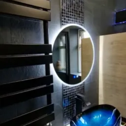 Nowoczesna łazienka z ciemnymi płytkami, podświetlanym okrągłym lustrem, dekoracyjną mozaiką, grzejnikiem drabinkowym i umywalką w kolorze niebieskim.