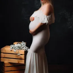 Kobieta w obcisłej, białej sukni z falbanami, w zaawansowanej ciąży, pozuje profilowo na ciemnym tle, trzymając dłonie na brzuchu; obok niej drewniane skrzynki z białymi kwiatami.