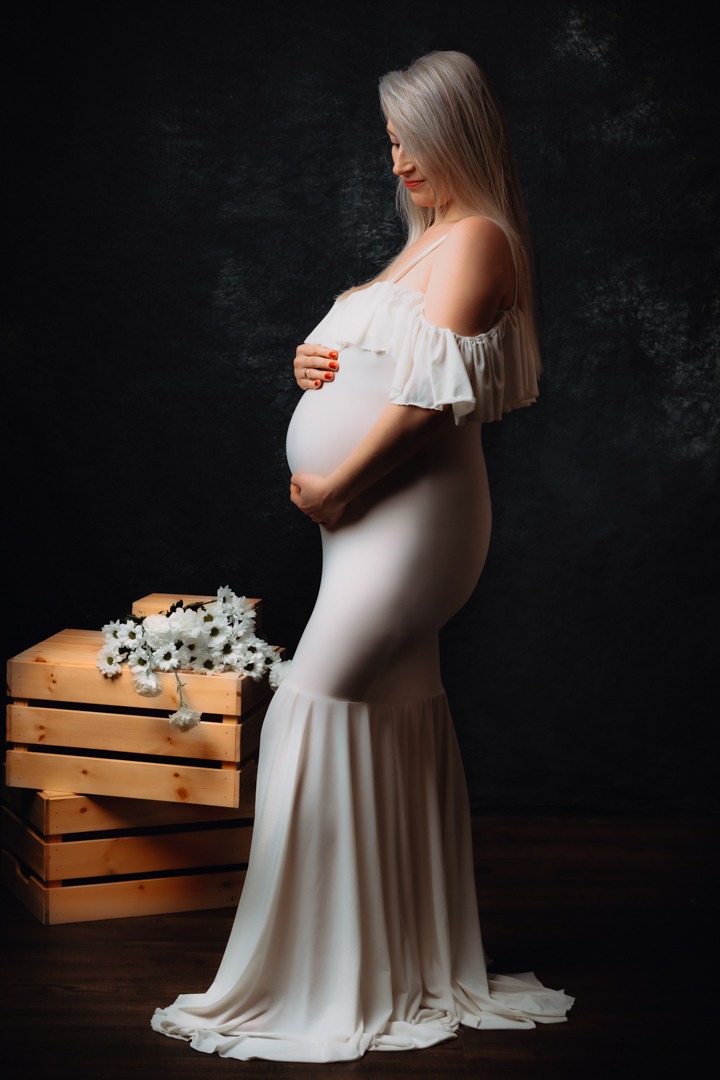 Kobieta w obcisłej, białej sukni z falbanami, w zaawansowanej ciąży, pozuje profilowo na ciemnym tle, trzymając dłonie na brzuchu; obok niej drewniane skrzynki z białymi kwiatami.