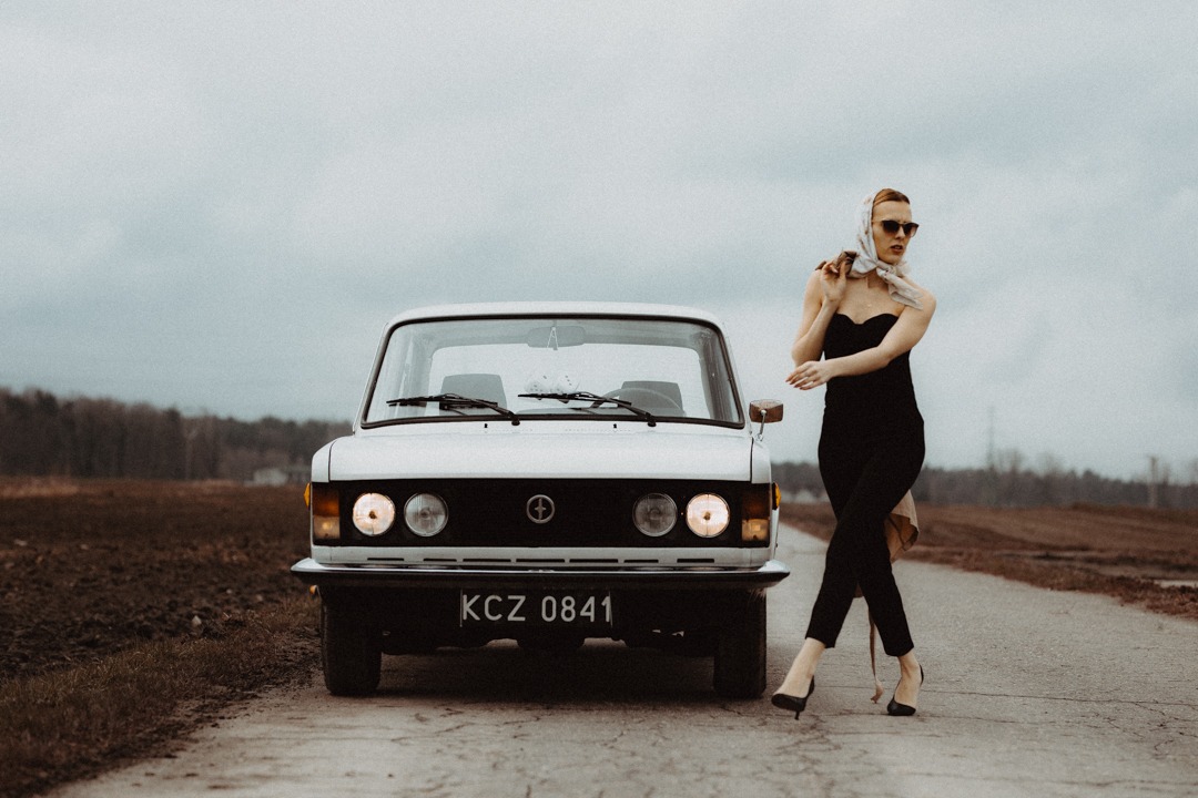 Elegancka kobieta w czarnym kombinezonie i apaszce pozuje obok białego, klasycznego Fiata 125p na polnej drodze w pochmurny dzień.