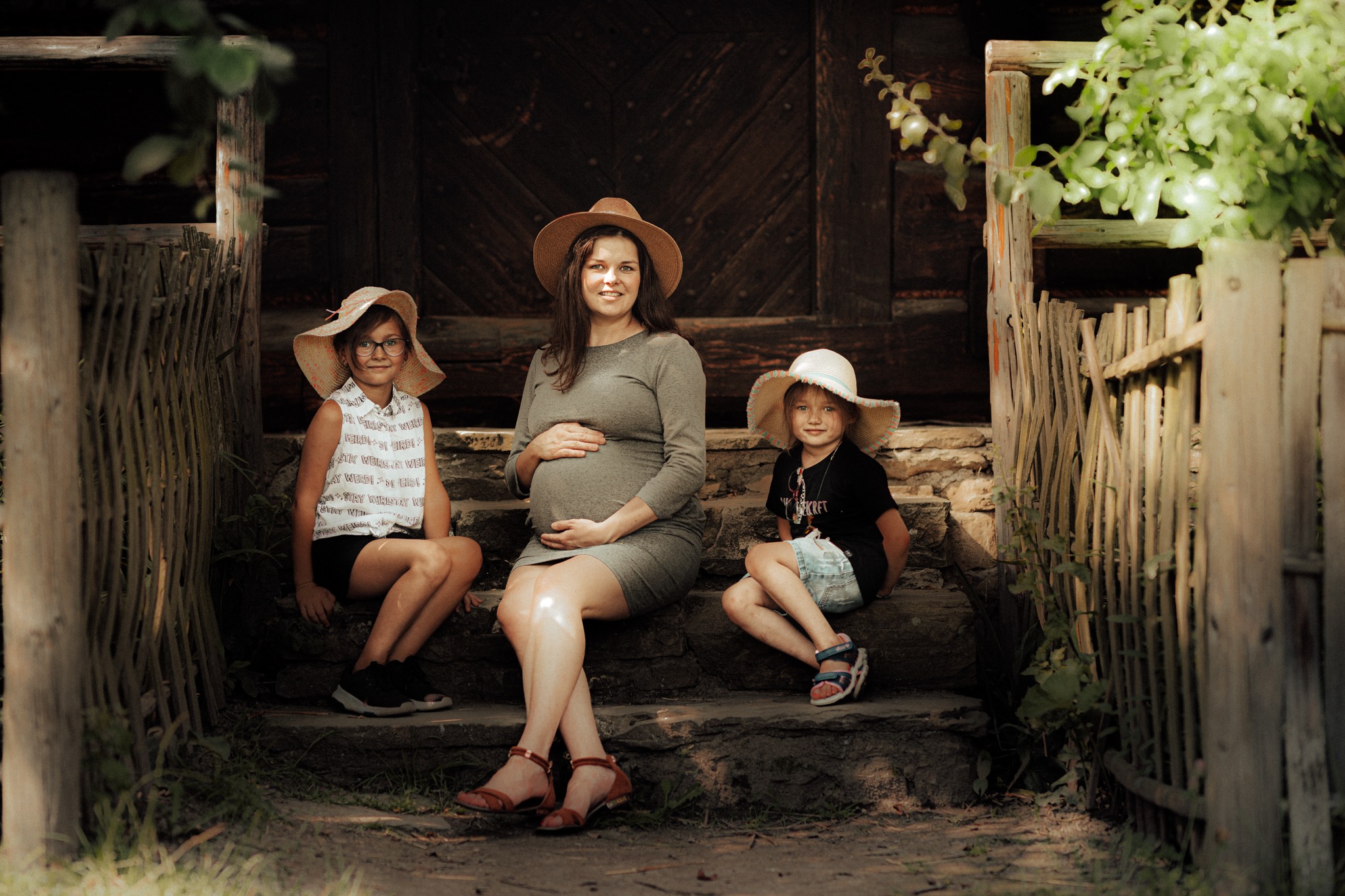 Przyszła mama w szarej sukience i kapeluszu siedzi na kamiennych schodach starej drewnianej chaty z dwiema dziewczynkami w kapeluszach, stylizowane ujęcie w rustykalnym klimacie.