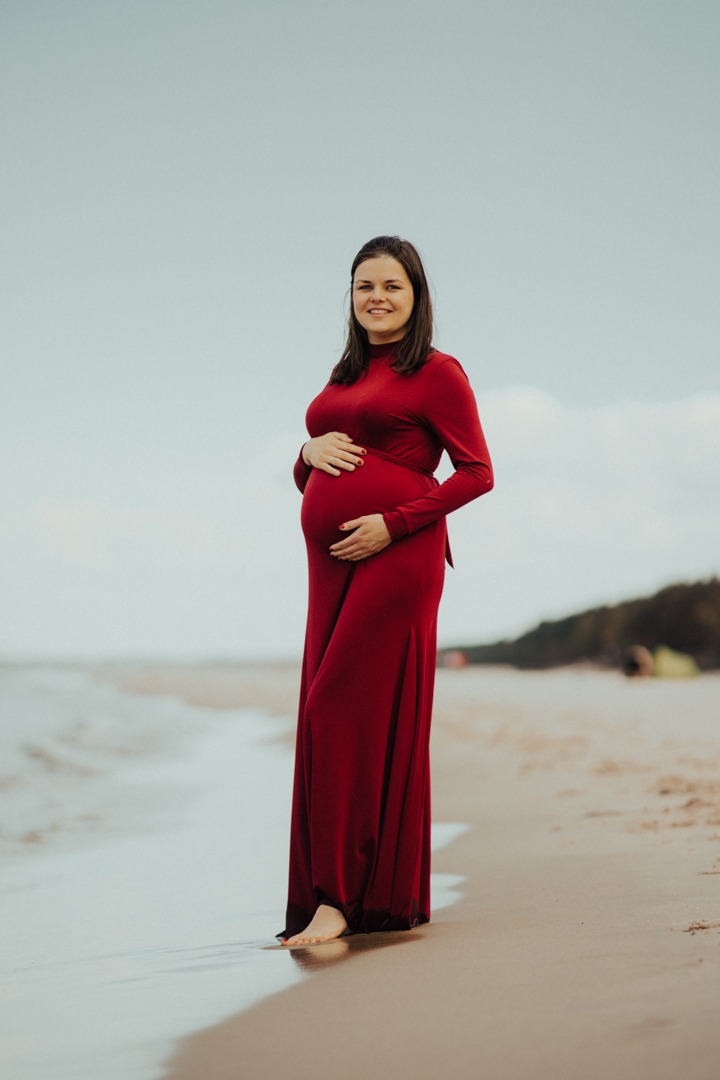 Kobieta w czerwonej, długiej sukni na plaży, trzymająca dłonie na brzuchu, z delikatnym uśmiechem.