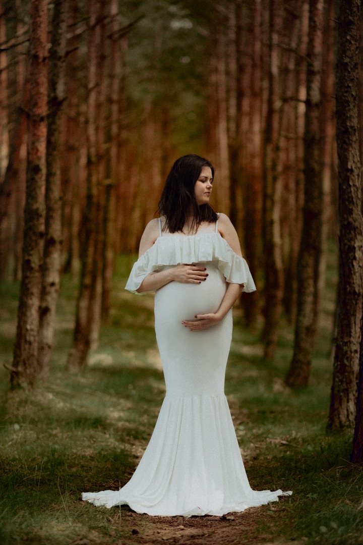 Kobieta w białej sukni ciążowej z falbankami, trzymająca ręce na brzuchu, pozuje w lesie z wysokimi drzewami.