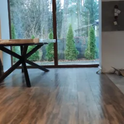 Wnętrze pokoju z brązową podłogą imitującą drewno, widok na ogród przez duże okno, fragment stołu z czarnymi nogami, nowoczesne lampy wiszące.