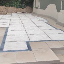 Wykończony taras z jasnych i ciemnych płytek, widoczne fragmenty elewacji budynku w odcieniach zieleni i beżu, w tle usypana pryzma ziemi.