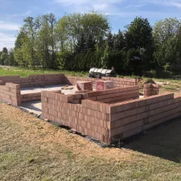 Wznoszone ściany domu z czerwonej cegły Porotherm, widoczne palety z cegłami, teren budowy w słoneczny dzień.