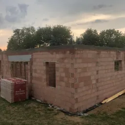 Budowa domu z czerwonej cegły Porotherm, widoczne pakiety cegieł na trawie, stan surowy otwarty, teren budowy.